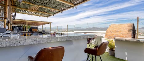 A rooftop bar with a stone counter, two brown bar stools, and a view of the mountains.