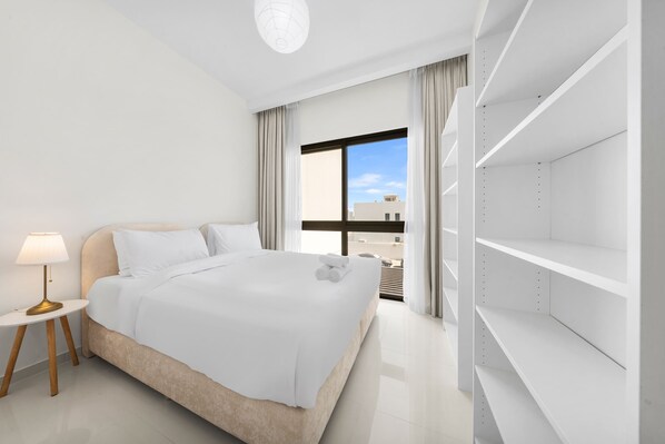 A modern hotel room with a large bed, a bedside table with a lamp, a view of the city through a large window, and built-in shelving. - Free Housekeeping Visits During Your Stay - Stayshort - Modern 4 BR Villa in Damac Hills 2 With Garden That Sleeps 8 (Dubai)