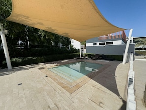 A swimming pool with a sunshade, outdoor seating, and a modern building in the background. - Free Housekeeping Visits During Your Stay - Stayshort - Modern 4 BR Villa in Damac Hills 2 With Garden That Sleeps 8 (Dubai)