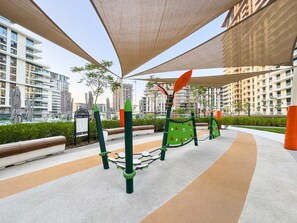 Ein Spielplatz im Freien mit Klettergerüsten und einer Sitzgelegenheit, umgeben von modernen Gebäuden und Grünflächen.