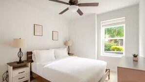 Una camera da letto con un letto, comodini, lampade, una ventola a soffitto, una finestra con vista e quadri appesi al muro.