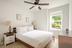 Ein Schlafzimmer mit Bett, Nachttischen, Lampen, Deckenventilator, Fenster mit Blick und gerahmten Bildern an der Wand.
