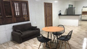 Een kamer met houten vloer, een bank, een ronde tafel met stoelen, en een keukenhoek met kastjes en apparaten.
