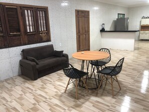 Una habitación con suelo de madera, un sofá, una mesa redonda con sillas y una zona de cocina con armarios y electrodomésticos.