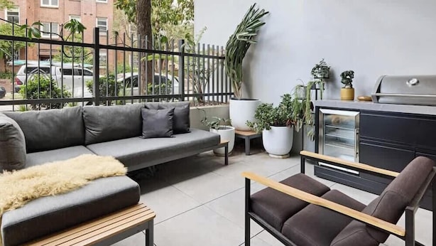 A modern outdoor living space with a grey sofa, a wooden bench, a black chair, a grill, and potted plants. - The Brass Banana in Petersham (Sydney)