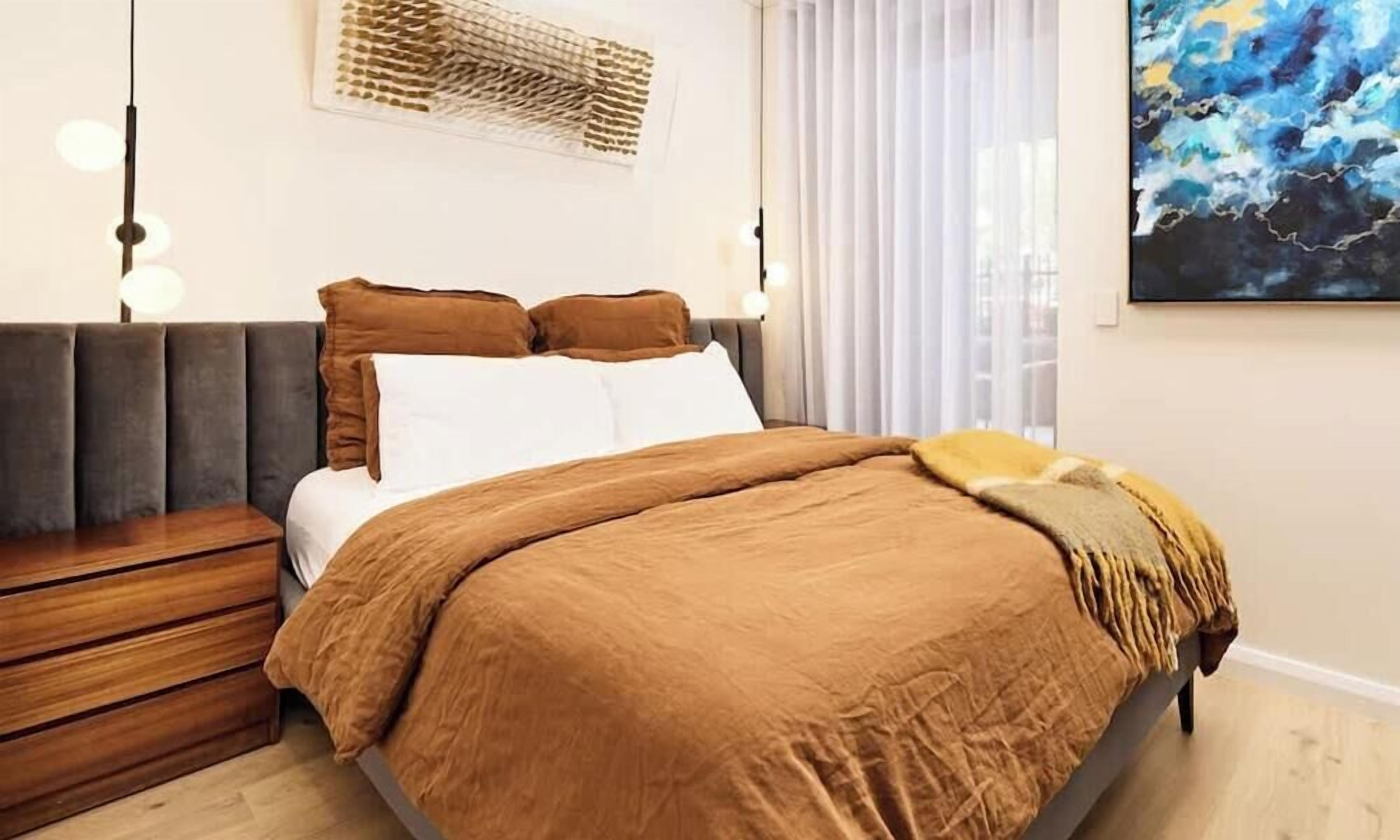 A bedroom with a bed, bedside tables, a wall-mounted light fixture, and a window with blinds.