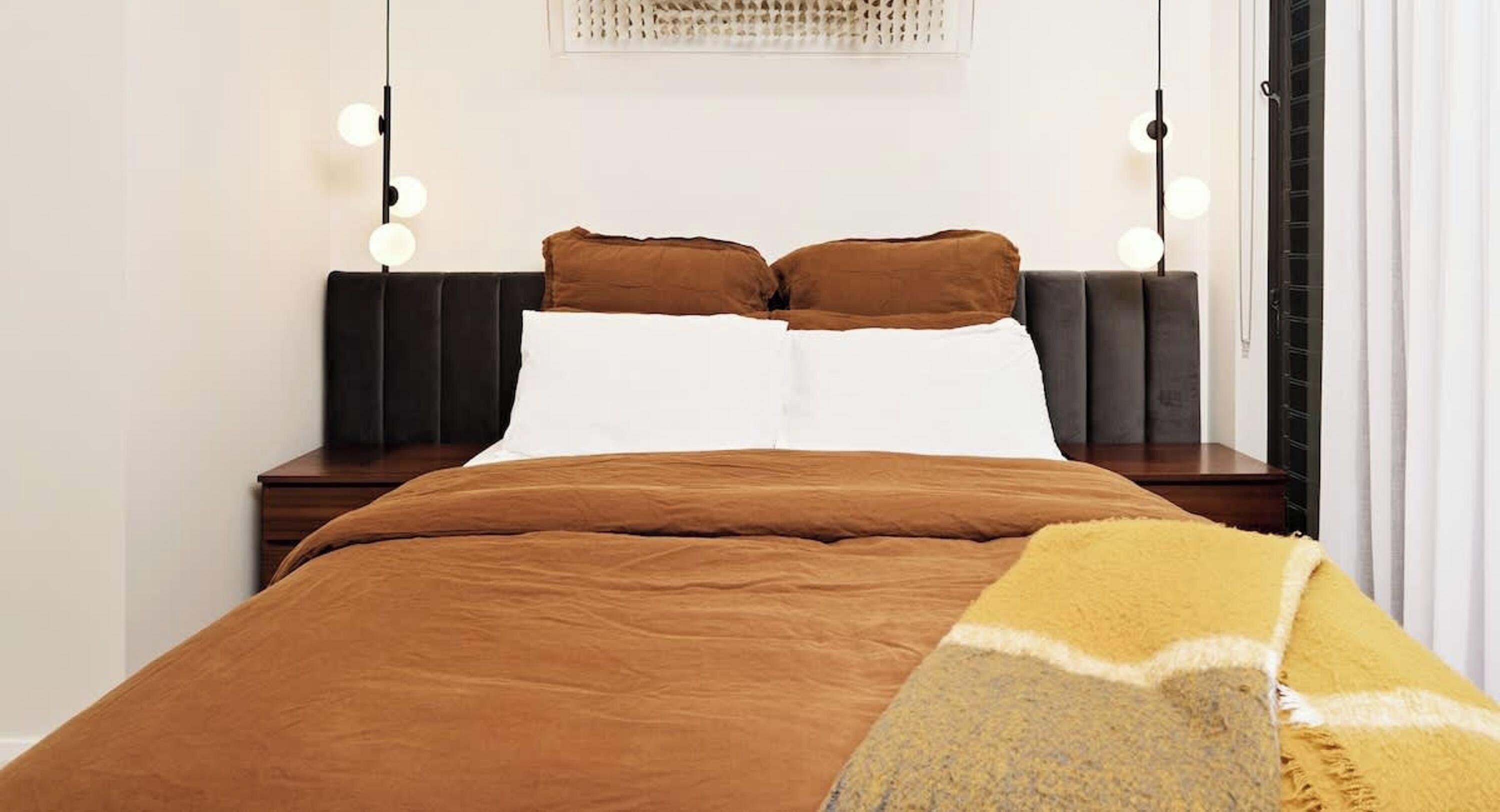 A neatly made bed with brown bedding, a white pillow, and a yellow throw blanket. There are two bedside tables with lamps and a wall-mounted light fixture.