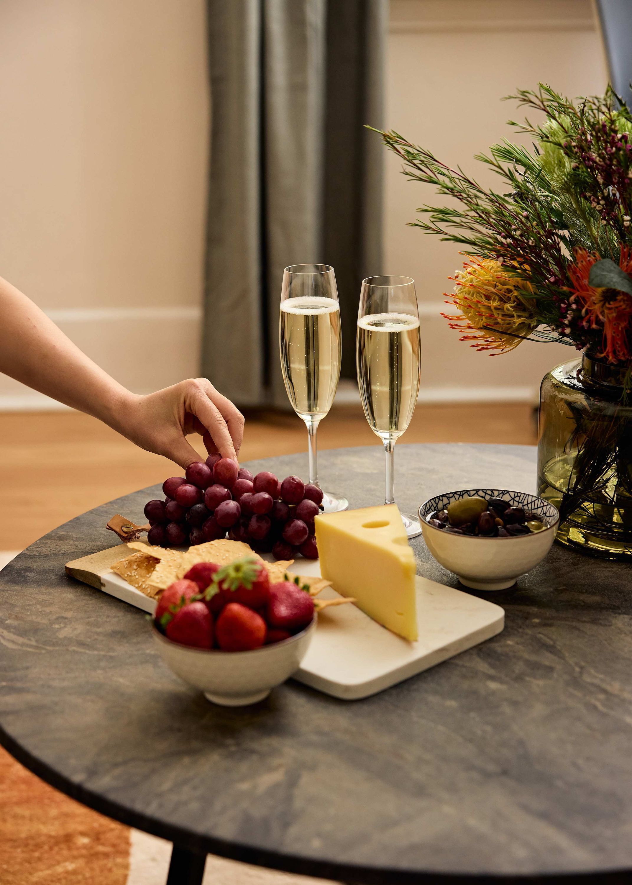 A round table with a cheese, grapes, strawberries, a bowl of olives, and two glasses of champagne.