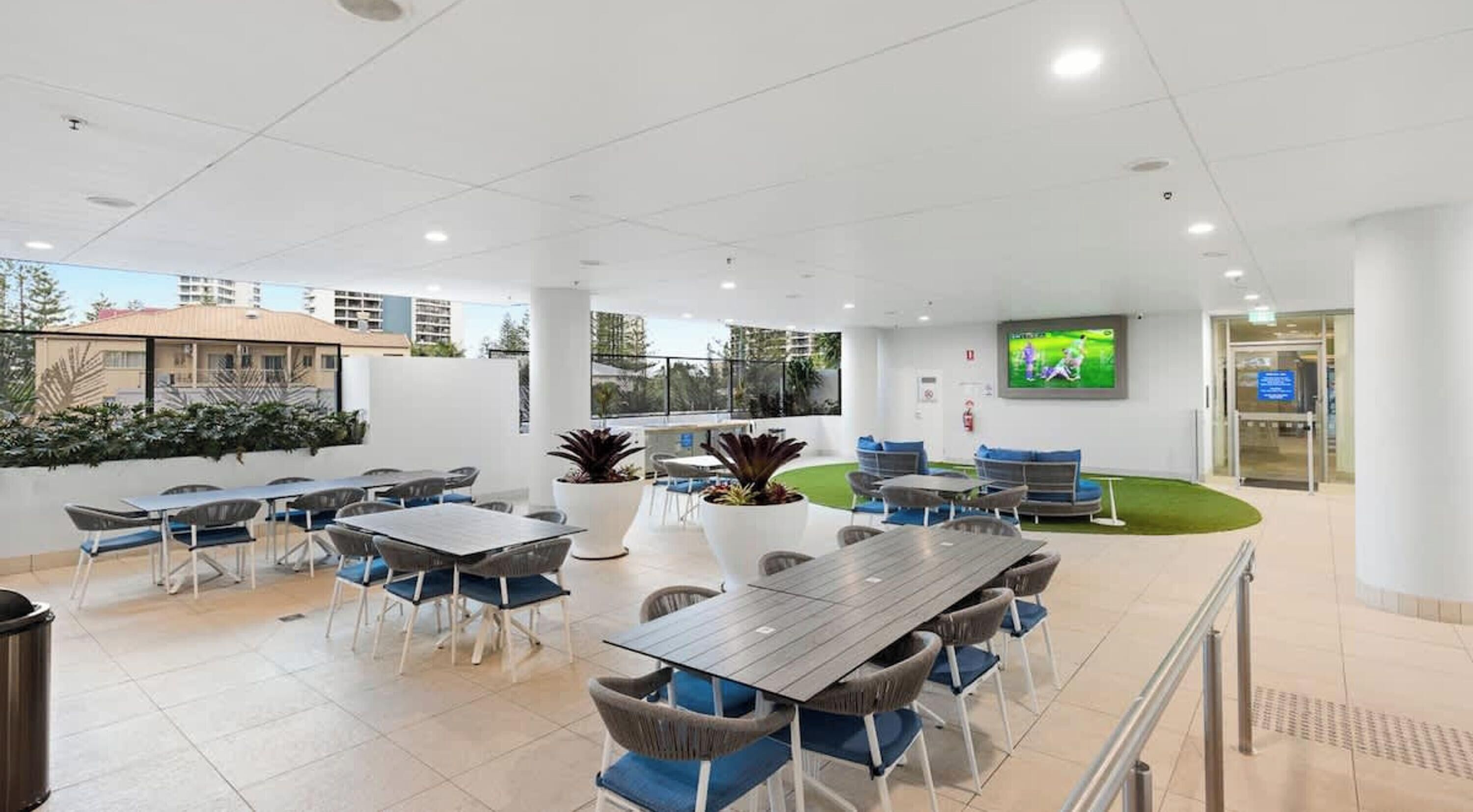 A spacious outdoor area with multiple tables and chairs, a flat-screen TV mounted on the wall, and a small grassy area.