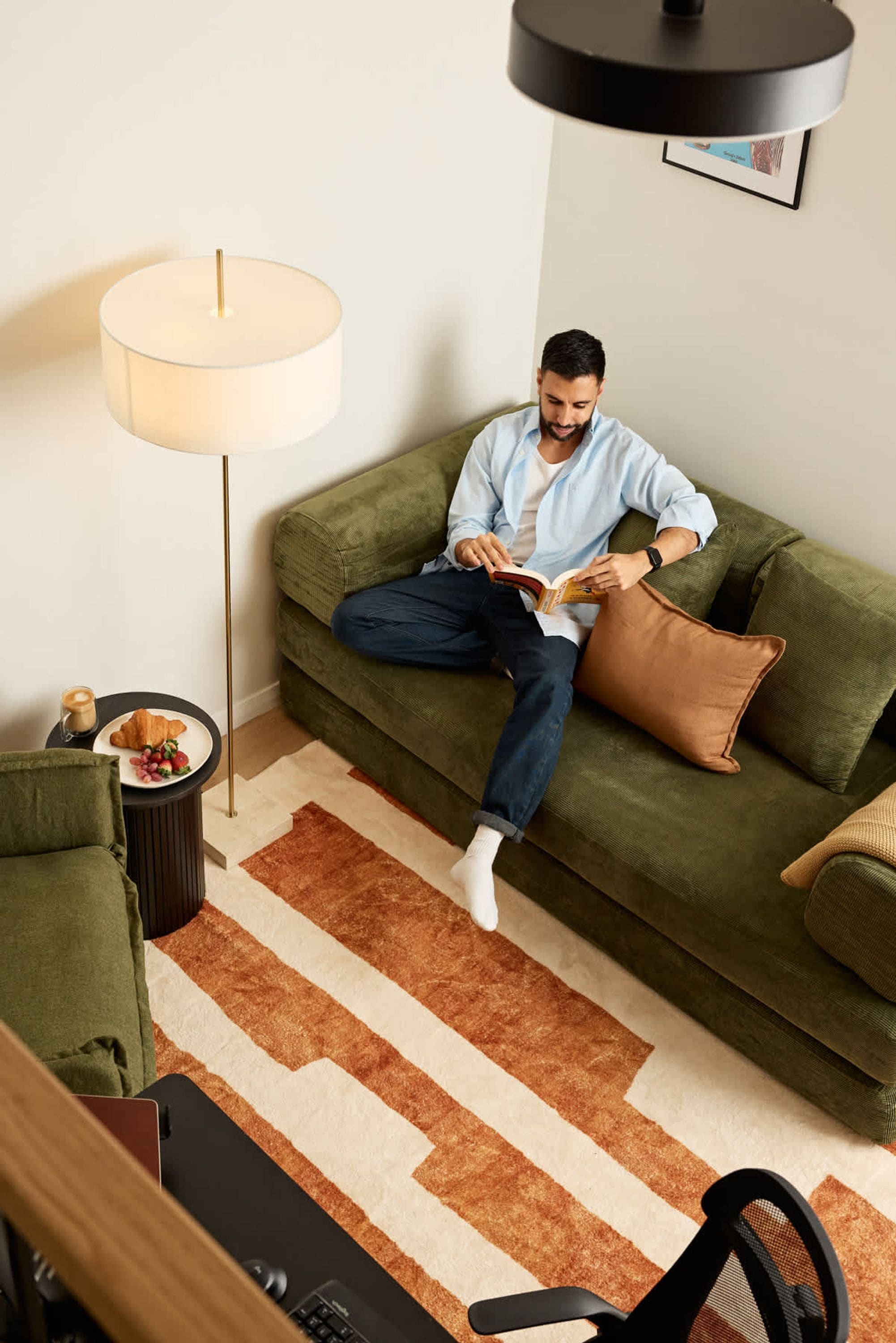 A person sitting on a green sofa reading a book, with a lamp and a small table with a croissant and coffee.