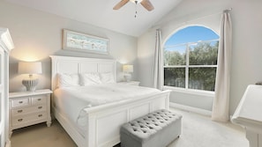 Quarto com uma cama grande, mesa de cabeceira, um banco, janela com cortinas e ventilador de teto.