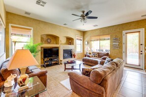 A living room with a sofa, a glass coffee table, a television, a fireplace, and a ceiling fan. - Private Pool & Hot Tub | Pet-Friendly Escape! (Maricopa)