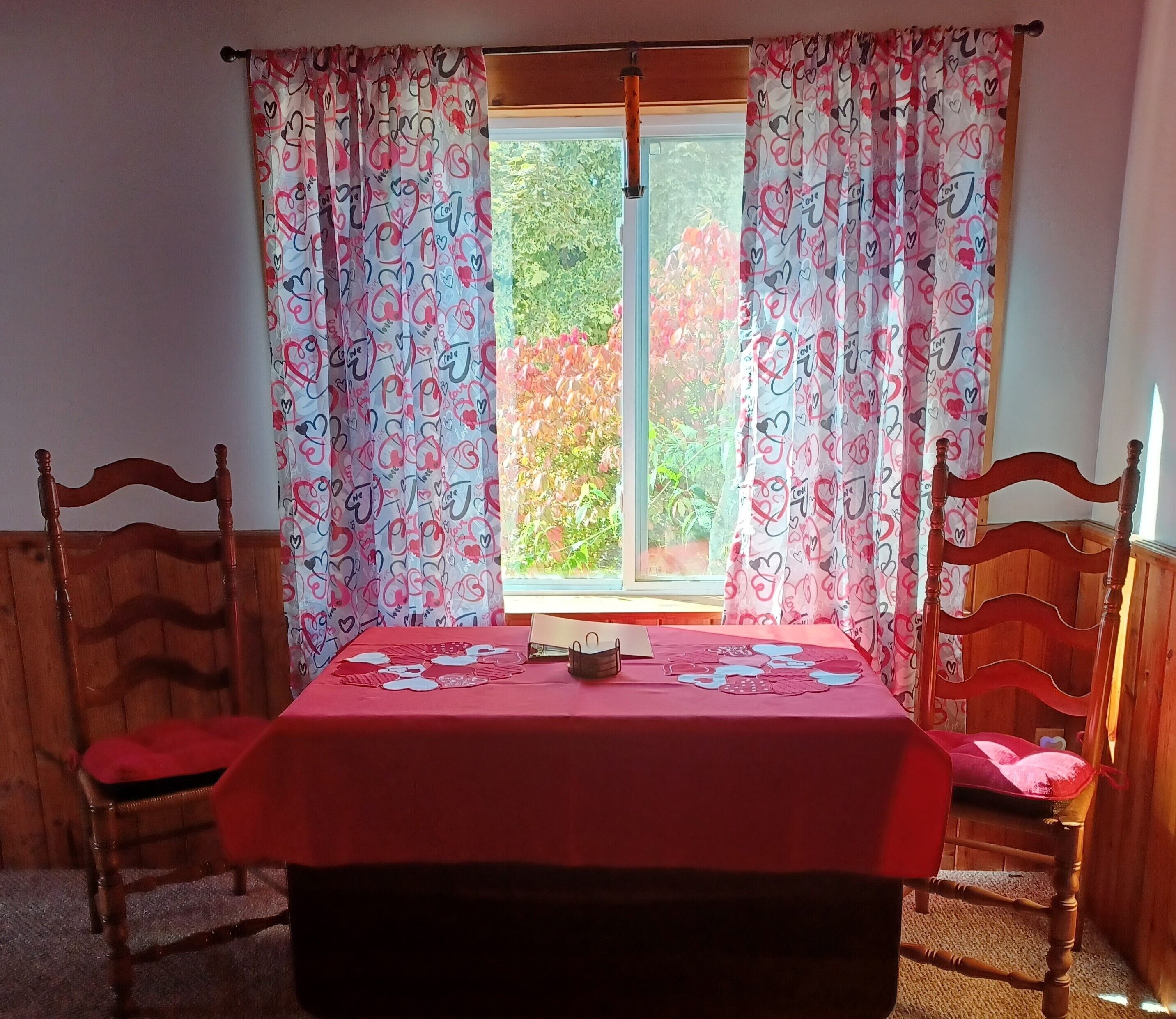 A room with a wooden table and chairs, a window with curtains, and a view of greenery outside.