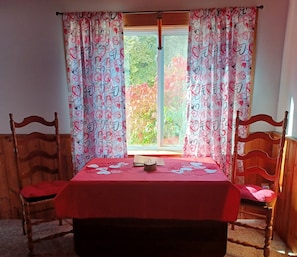 Une pièce avec une table et des chaises en bois, une fenêtre avec des rideaux et une vue sur la verdure à l’extérieur.