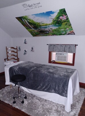 A bedroom with a bed, a chair, a window with a curtain, a wall with a painting, and a thermostat.