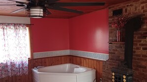 A bathroom with a bathtub, a wooden ceiling, a red wall, a ceiling fan, and a brick wall with a stove. - Romantic couples bed & breakfast in secluded Strykersville NY (STRYKERSVILLE)