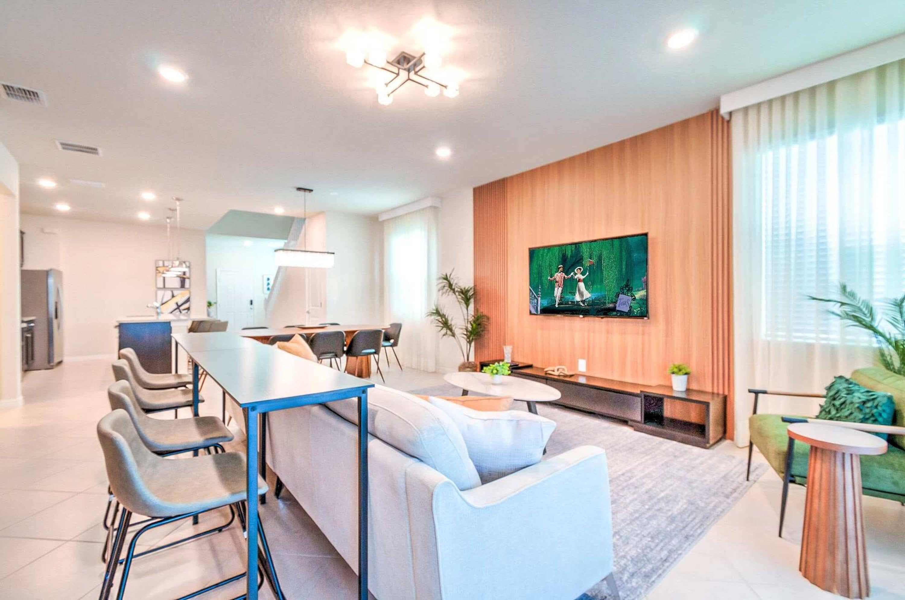 A modern living room with a sofa, dining table, chairs, a TV, and a kitchen area in the background.