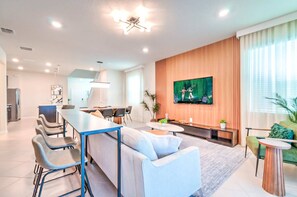 Ein modernes Wohnzimmer mit einer Couch, einem Esstisch, Stühlen, einem Fernseher und einer Küche im Hintergrund.