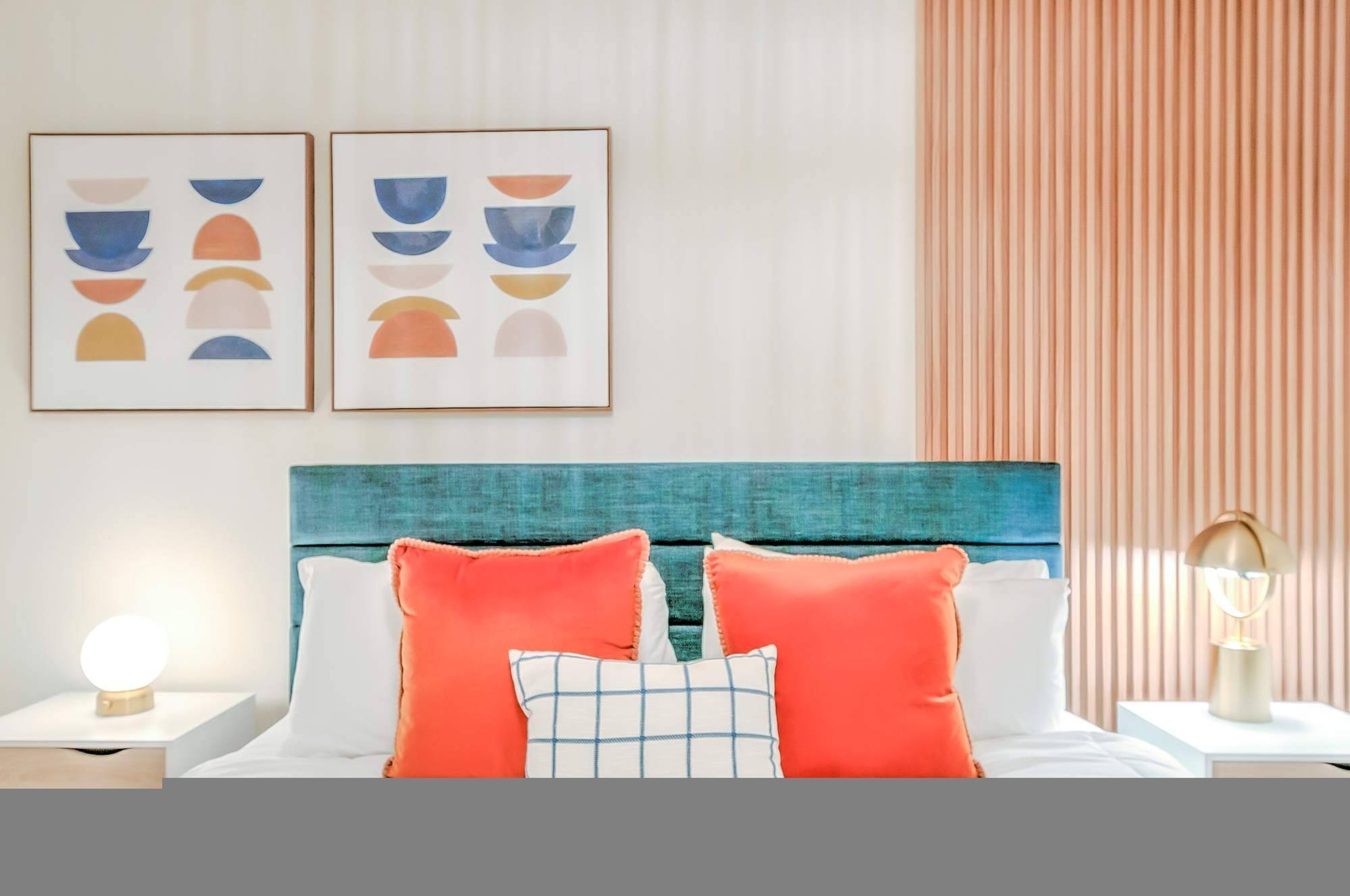 A neatly made bed with a teal headboard, two framed abstract paintings, and a bedside lamp.
