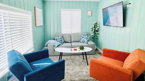 A room with teal walls, a grey sofa, blue and orange chairs, a round coffee table, and a mounted TV. - Charming Beach Cottage (San Diego)