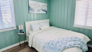 A bedroom with a bed, a nightstand, a lamp, and a wall-mounted artwork. - Charming Beach Cottage (San Diego)