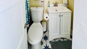 A bathroom with a toilet, sink, mirror, and a window with blinds. - Charming Beach Cottage (San Diego)