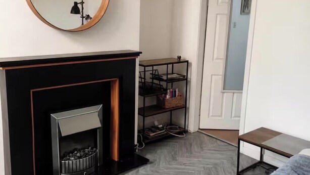 A living room with a fireplace, a round mirror, a grey sofa, a small table, and a chandelier. - Park4Free - Etihad & Co-op Live near- Nifty Find (Manchester)