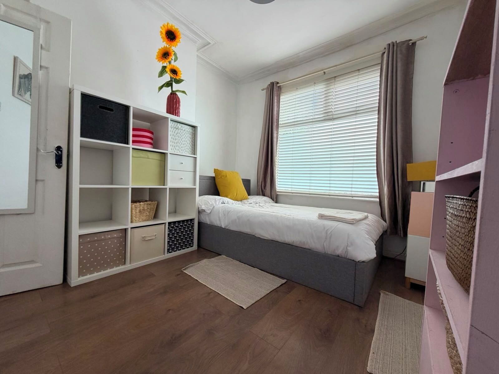 A bedroom with a bed, a wardrobe, a shelf unit, a window with curtains, and a vase with sunflowers.. Park4Free - Etihad & Co-op Live near- Nifty Find