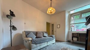 A modern living room with a sofa, a floor lamp, a chandelier, a table, a rug, and a window with blinds. - Park4Free - Etihad & Co-op Live near- Nifty Find (Manchester)