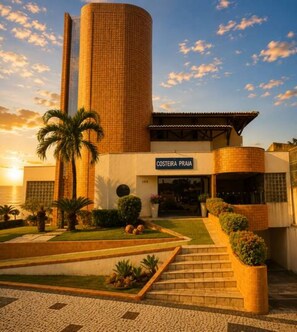 A hotel with a prominent cylindrical tower, a sign reading 'COSTEIRA PRAJA,' and a well-maintained garden with palm trees and shrubs.
