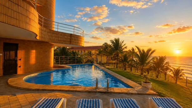 En swimmingpool med liggestole, en bygning med balkon og udsigt til solnedgang.