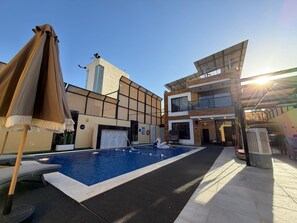Une piscine avec des chaises longues, un espace abrité et un bâtiment au design moderne, doté d’une tour.