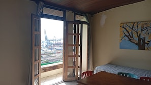 A room with a wooden table, chairs, a painting of a tree, and a view of a port with cranes. - Direita S.Antonio Direita S.Antonio -Charmant appartement baie de Todos Santos. (Salvador)