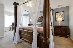 A four-poster bed with a canopy, a bedside table with a lamp, a framed artwork on the wall, and a view of the room's interior. - Happy Planet 1: Private Pool and Spa! BBQ Grill. (Davenport)