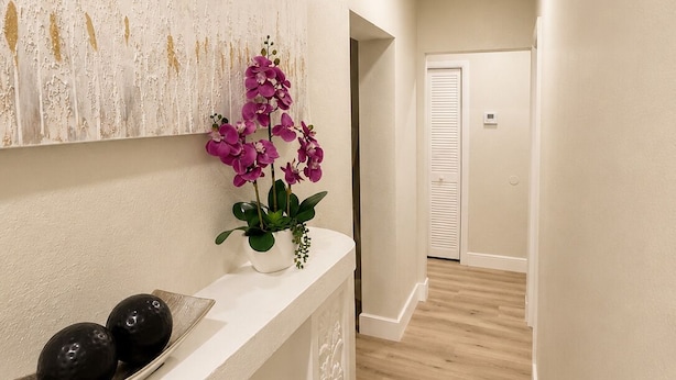 Um corredor com parede branca, um painel decorativo com detalhes dourados, um console branco com um vaso de flores roxas e uma tigela preta.