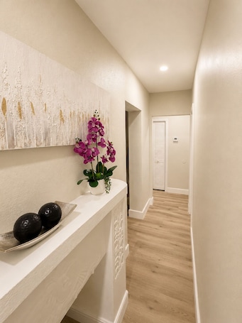 A hallway with a white wall, a decorative panel with gold accents, a white console table with a vase of purple flowers, and a black bowl.