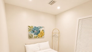 Quarto com cama, mesa de cabeceira, luminária pendente, janela com persianas e um respiradouro na parede.