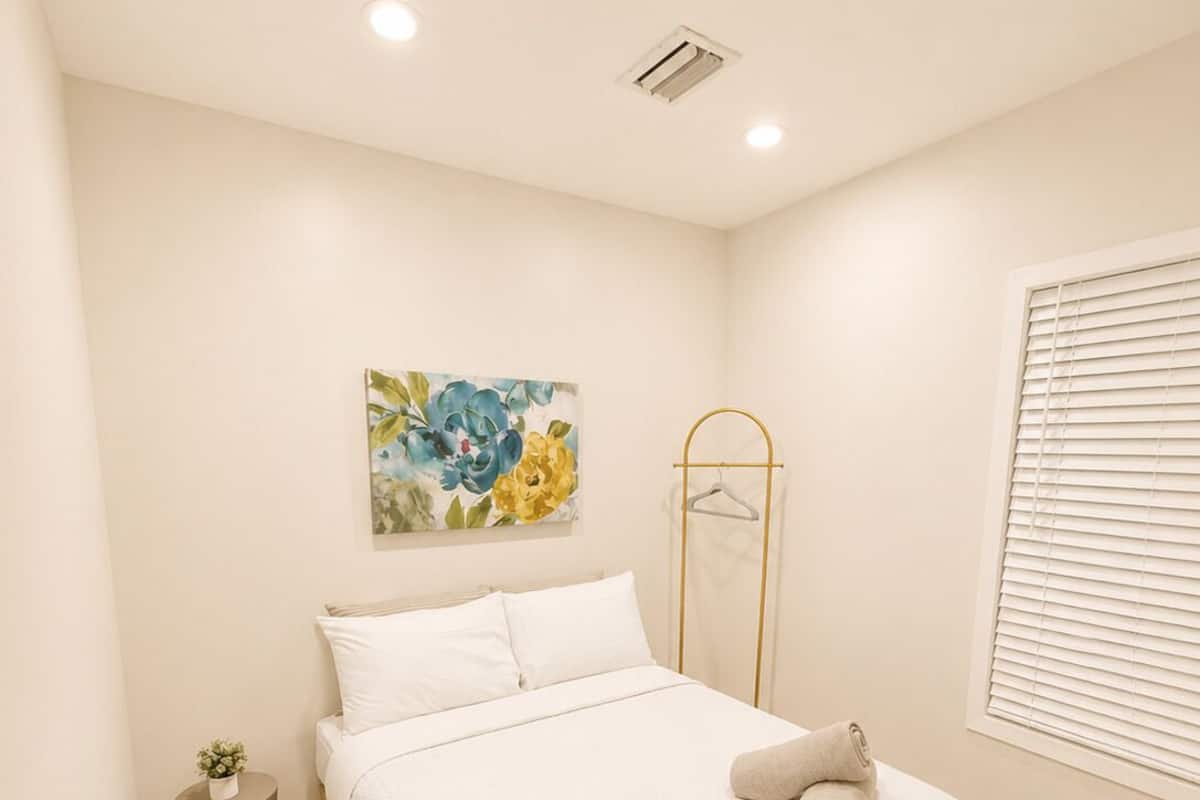 A bedroom with a bed, a bedside table, a hanging light, a window with blinds, and a wall-mounted air vent.