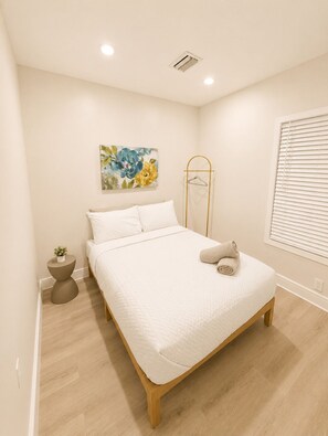 A bedroom with a bed, a bedside table, a hanging light, a window with blinds, and a wall-mounted air vent. - Villa Shores  (Miami)