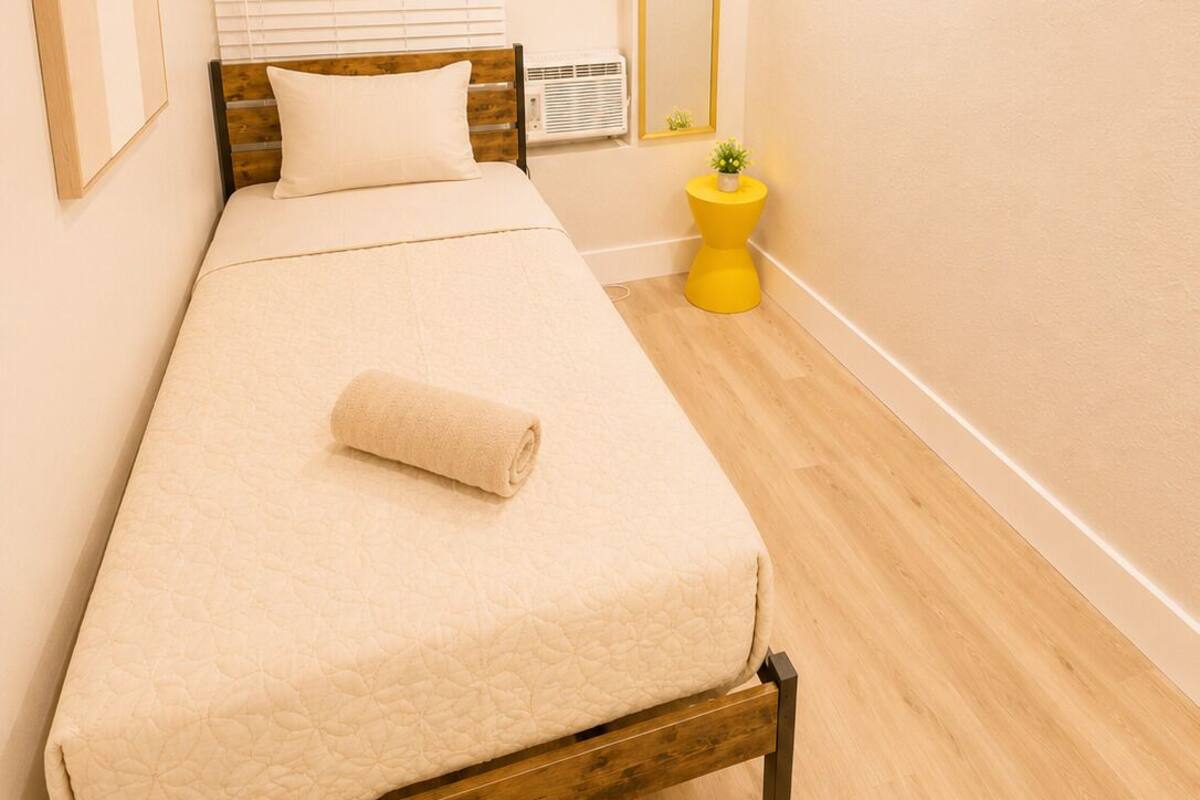 A bedroom with a bed, a bedside table, a window with blinds, a mirror, and a small plant.