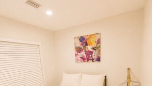 Quarto com cama, janela com persianas, ar condicionado fixado na parede e um quadro abstrato colorido.