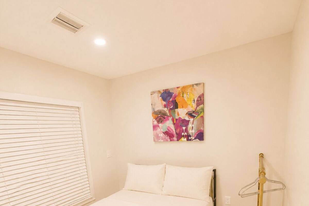 A bedroom with a bed, a window with blinds, a wall-mounted air conditioner, and a colorful abstract painting.
