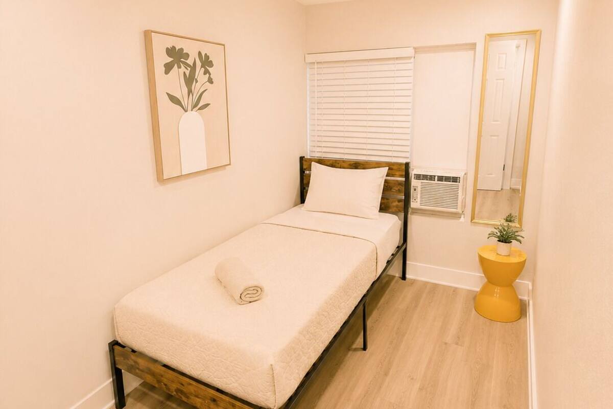 A small, neatly arranged bedroom with a single bed, a bedside table with a plant, a window with blinds, and a mirror on the wall.