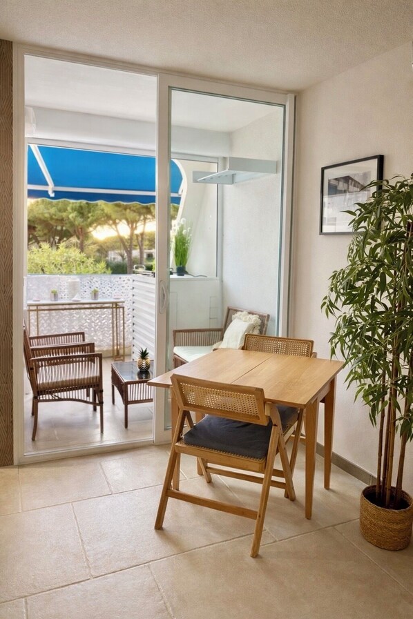 Une pièce avec une table et des chaises en bois, une plante en pot et une porte-fenêtre coulissante donnant sur un espace extérieur doté d’un auvent bleu.