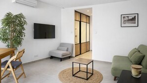 Una sala moderna con un televisor de pantalla plana, un sofá verde, una mesa de comedor de madera con sillas, una mesita auxiliar y una planta grande en maceta.