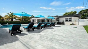 Un espace aménagé au bord de la piscine, avec des chaises longues et des parasols, une piscine, et un bâtiment doté de grandes fenêtres.