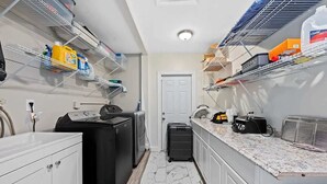 Une cuisine de taille réduite, équipée d’un lave-linge, d’un réfrigérateur compact et d’un plan de travail sur lequel sont disposés divers objets.