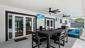 Une terrasse extérieure avec une table et des chaises, un canapé, des ventilateurs de plafond et une porte-fenêtre coulissante donnant sur une zone avec piscine.