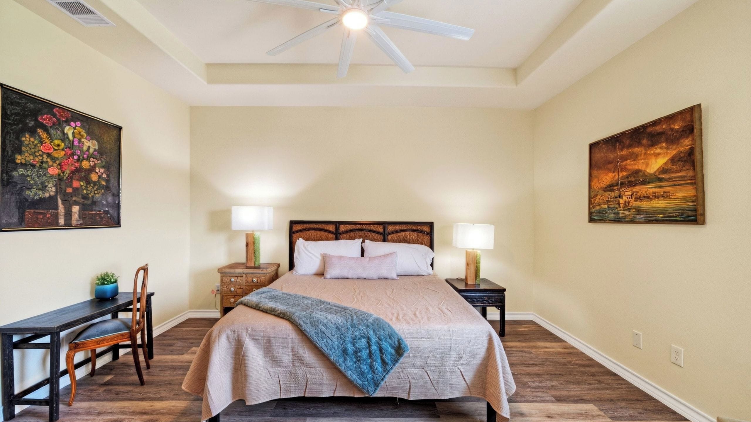 Quarto com cama, duas mesas de cabeceira, uma cadeira, ventilador de teto e duas pinturas nas paredes.