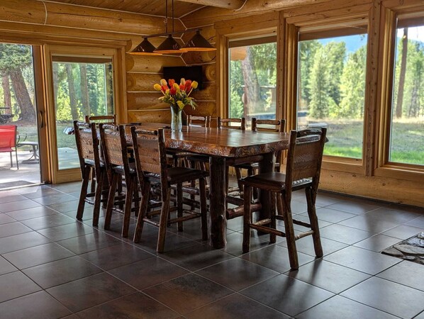 A wooden dining table with chairs, a vase of flowers, and large windows with a view of trees.